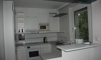 A modern, bright kitchen with white cabinets and a minimalist design. The countertop is made of light stone and a large window provides daylight.