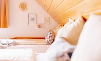 A cozy bedroom with wooden walls and bright cushions. Decorative pictures hang on the wall and the windows are furnished with curtains.