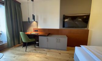 A modern room with a television, a desk, and a comfortable chair. The wooden floors and large windows create a bright atmosphere.