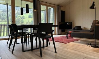 A modern living room with a dining table and black chairs. Large windows offer a view of the green landscape.