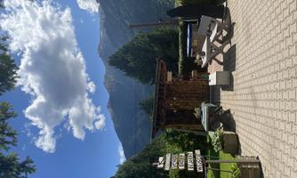 A cozy terrace with benches and tables, surrounded by trees and mountains. The sky is clear with a few clouds.