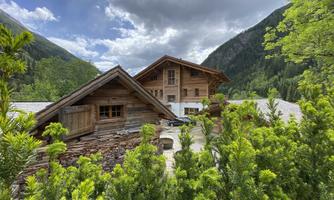 A rustic wooden cabin surrounded by green trees and mountains. The sky is partly cloudy with some rays of light.
