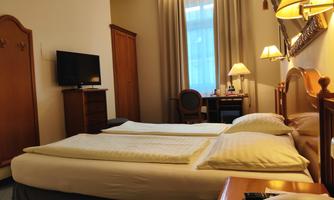 A comfortable hotel room with two single beds and a small desk. The room features a TV and a large window with curtains.