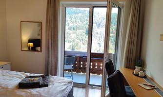 A cozy room with a bed, a desk, and a balcony. Through the window, you can see a beautiful view of the surrounding mountains.
