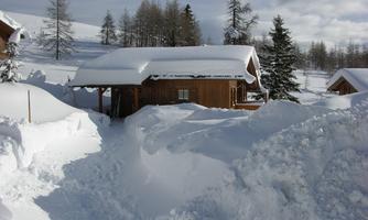 A wooden cabin in a snowy landscape. Deep snow surrounds the house and trees are visible in the background.