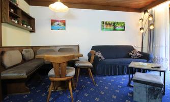 A cozy living room with a blue upholstered sofa and a dining table. Pictures of natural motifs hang on the wall, and the floor is covered with a blue carpet.
