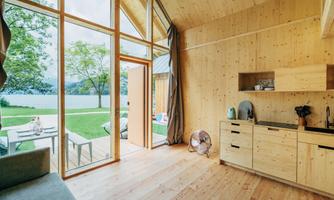A modern wooden house with large windows overlooking a lake. Inside, there is a kitchenette and a cozy living area.