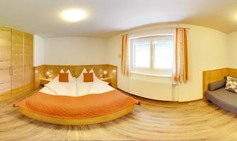 A cozy bedroom with a large bed and orange pillows. There is a wooden wardrobe and a gray sofa next to a window.