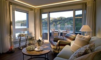 A stylish living room with a view of the water and the mountains. The decor is modern and cozy, featuring a table and comfortable seating.