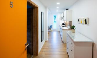 A bright hallway with an orange door and modern furniture. At the end of the hallway, there is a bright kitchen with plants and pictures on the wall.