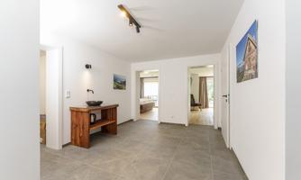 A bright, modern entrance area with gray tiles and a wooden table. In the background, there are additional rooms and windows that provide plenty of natural light.