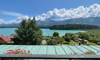 A picturesque landscape with a clear blue lake and mountains in the background. In the foreground, plants are blooming on a balcony.