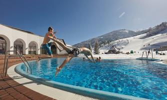 A boy jumps into a pool while another watches. Snow-covered mountains can be seen in the background.