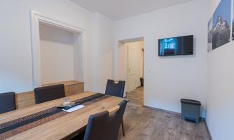 A modern room with a large wooden table and black chairs. A television hangs on the wall, and in the background, a door is visible.