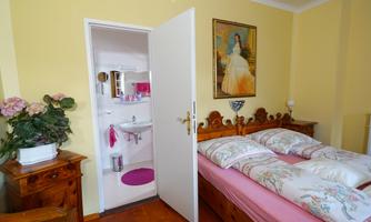 A cozy bedroom with a double bed and a wooden wardrobe. A bathroom is visible through the door, with a fresh, bright atmosphere.