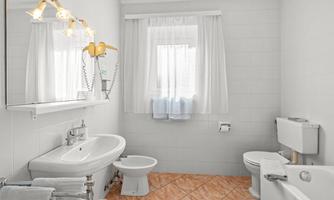 A modern bathroom with a bright tiled floor and white walls. It features a sink, a toilet, and a window with curtains.