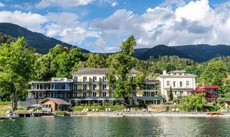 A picturesque shore with several hotels and lush greenery. In the background, gentle mountains rise under a blue sky with white clouds.