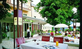 A cozy café with outdoor seating under shade-giving trees. Guests enjoy their drinks at tables while a waitress serves.