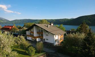 A beautiful house by the shore of a lake, surrounded by green trees and mountains. The sky is blue with a few clouds.