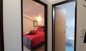 A cozy bedroom with red bedding and modern pillows. In the background, a bathroom can be seen.
