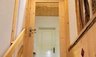 A narrow hallway with a wooden staircase and a glass door. The walls and the wooden railing are made of light wood.