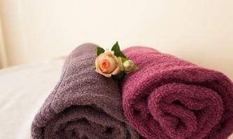 Two rolled-up towels in purple and pink are lying on a bed. A small pink rose and some leaves are decorated between the towels.