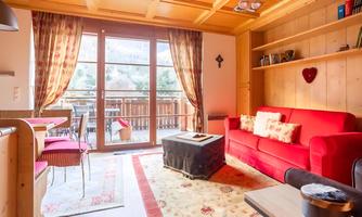 A cozy living room with a red sofa and wooden elements. Large windows offer a view of the terrace and the surroundings.
