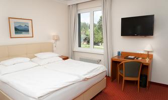 A cozy hotel room with a double bed and a desk. A window offers a view of nature and a television is mounted on the wall.