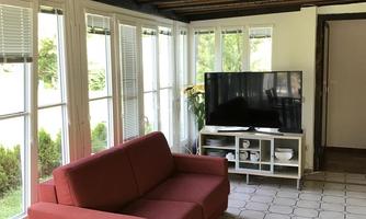A bright living room with large windows and a red sofa. A television is placed on a white TV unit in the corner.