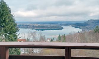 A view of a calm lake, surrounded by trees and gentle hills. The sky is cloudy, and the landscape appears peaceful.
