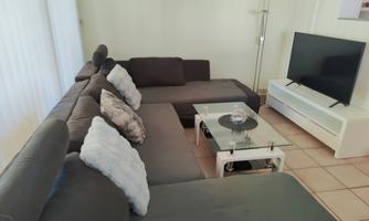 A cozy living room with a large couch and a modern television. A glass table stands in the center of the room.