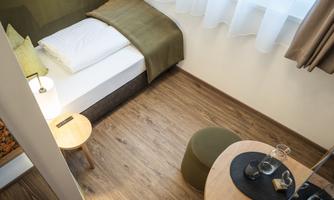 A modern, cozy room with a bed, a small table, and a chair. The floor is wooden and the room has large windows with plenty of natural light.
