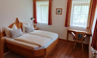 A cozy bedroom with a double bed and wooden details. The windows are bright and friendly, surrounded by fresh curtains.