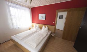 A cozy bedroom with two single beds and a red wall. The room has large windows, plenty of natural light, and a simple, warm style.
