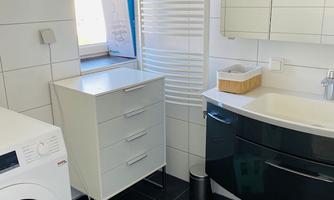 A modern bathroom with a washing machine, a white cabinet, and an elegant sink. Large windows let in plenty of light, and the walls are decorated with white tiles.