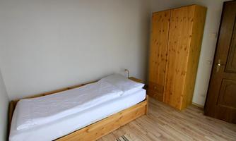 A simple and bright bedroom with a single bed and a wooden wardrobe. The floor is made of wood, and the walls are kept neutral.