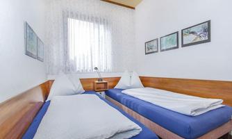 A simple room with two single beds and natural light sources. The walls are decorated with pictures, and there is a small table with a lamp.