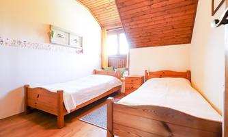 The image shows a cozy room with two bright wooden single beds. The walls are painted in soft colors, and a window provides plenty of daylight.