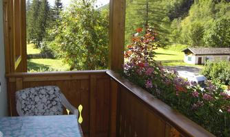 A cozy balcony with a table and a chair. In the background, green meadows and trees can be seen.
