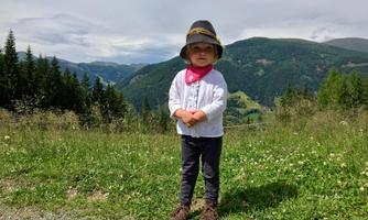 A small child with a sun hat and neck scarf is standing on a meadow. In the background, green mountains and a cloudy sky are visible.