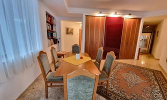 A cozy dining area with a wooden table and chairs. In the background, there are bookshelves and a carpet.