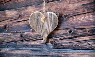A wooden heart hangs on a string in front of a rustic wooden wall. The natural texture of the wood gives the image a warm and inviting character.