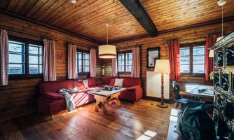 A cozy living room with wooden walls and a red sofa. The room has large windows and is brightly lit.