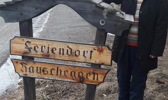 A woman stands next to a wooden signpost. The sign shows the name "Schweindorf Hausrachen" and is located in a forest environment.