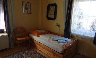 A cozy room with a single bed and a wooden chair. The walls are painted in warm colors and there are windows with curtains that let in daylight.