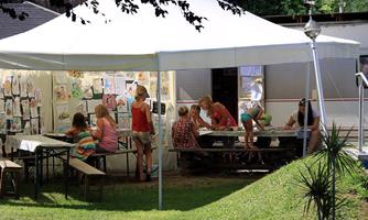 A tent with children working creatively and painting. Surrounded by a green meadow and pictures on the walls.