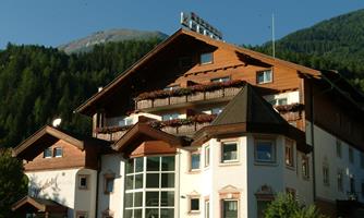 A cozy hotel building in Alpine style with wooden elements and balconies. In the background, green mountains and a clear sky can be seen.