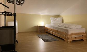 A simple, bright room with a bed, a nightstand, and a small rug. In the background, there is a clothing rack and a refrigerator.
