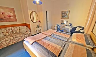 A cozy bedroom with two beds, light colors, and a sofa. Pictures hang on the wall, and a table with chairs is also available.