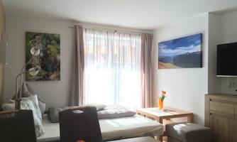 A bright living room with large windows and a view of the mountains. There is a cozy couch, a table, and decorative pictures on the walls.